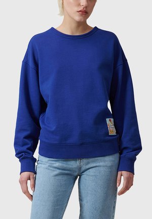 Persona che indossa una felpa blu royal semplice con una piccola toppa colorata vicino all'orlo e jeans azzurri, in piedi davanti a uno sfondo neutro.