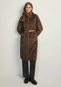 Brązowy płaszcz ze sztucznego futra z wzorem warkocza, wysokim kołnierzem i brązowym paskiem. Posiada przednie kieszenie i sięga do kolan. Czarne spodnie i buty.