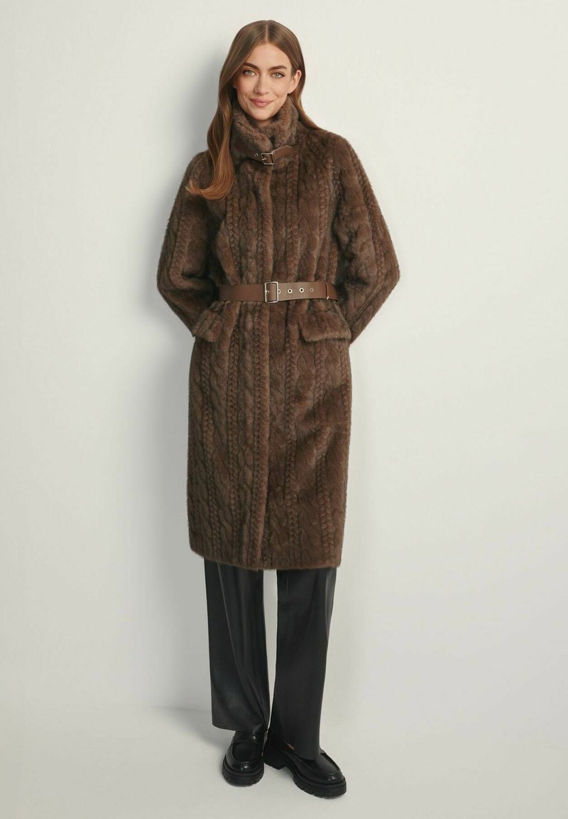 Brązowy płaszcz ze sztucznego futra z wzorem warkocza, wysokim kołnierzem i brązowym paskiem. Posiada przednie kieszenie i sięga do kolan. Czarne spodnie i buty.