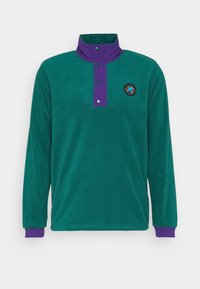 Tealfärgad fleecetröja med lila tryckknappskrage och muddar. Har en rund logga på bröstet. Mjuk textur och långa ärmar.