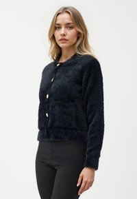 Cardigan nero peloso con scollatura rotonda, bottoni dorati e due tasche frontali. Design accorciato con maniche lunghe e una texture morbida.