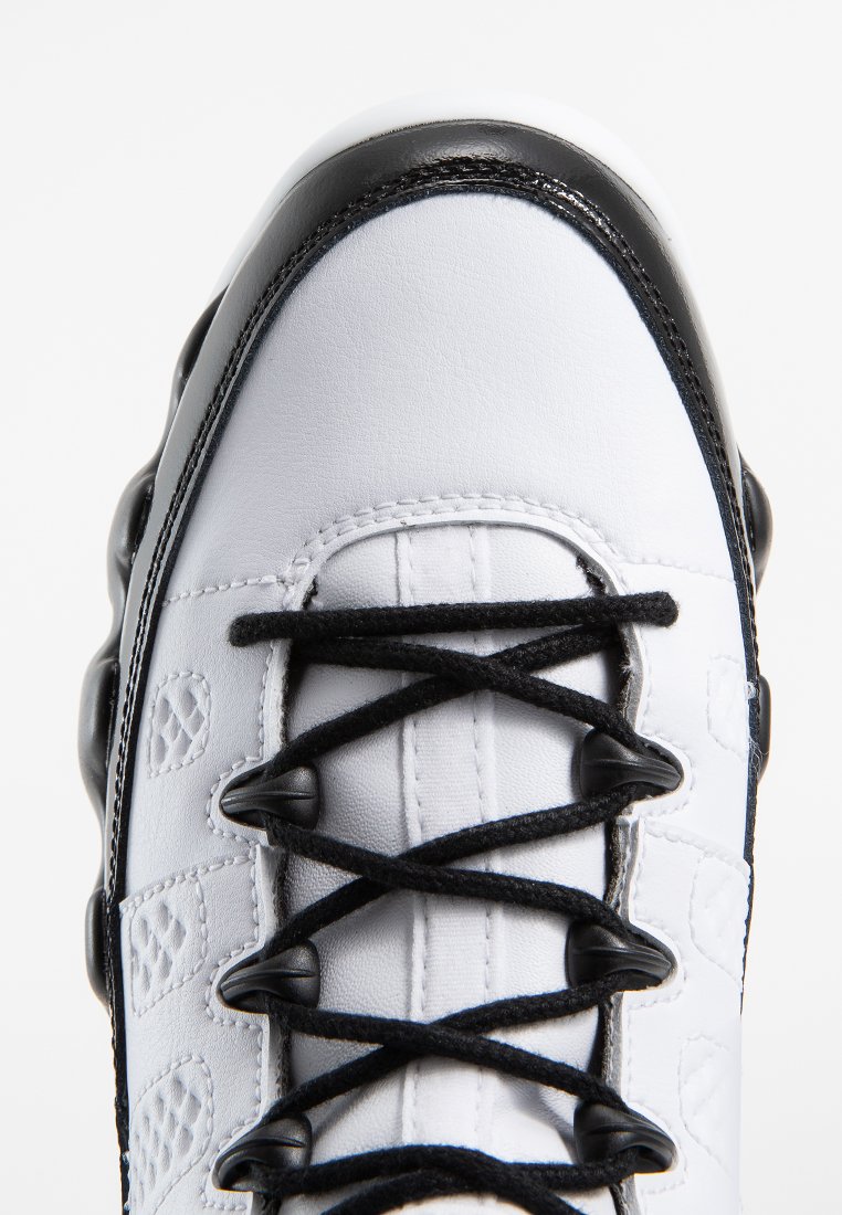 Białe sneakersy z czarnymi akcentami, gładki materiał syntetyczny, okrągły czubek i wyraźny system sznurowania. Widoczny teksturowany wzór po bokach.