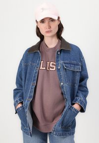 Denim jakna s rjavim ovratnikom, svetlo vijolična majica s kapuco z napisom 'Juicy' v debelem tisku in roza bejzbol kapa. Opuščen outfit s klasičnim slogom.