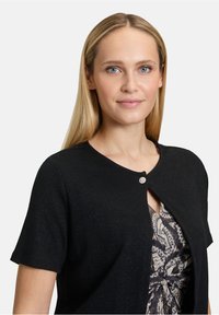 Schwarze kurzärmlige Strickjacke mit Rundhalsausschnitt und einem dekorativen silbernen Knopf auf der Vorderseite. Der Stoff hat einen dezenten Glanz.