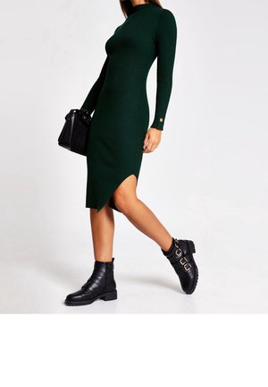 Robe en tricot côtelé verde avec un col montant, des manches longues et un ourlet asymétrique. Associée à des bottines noires à la cheville avec plusieurs sangles et éléments métalliques.