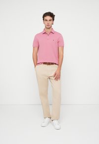 Rožinė polo maikutė su apykakle ir trimis mygtukais, derinama su beige kelnėmis ir baltomis sportinėmis batelėmis. Išskirtinis logotipas krūtinėje.