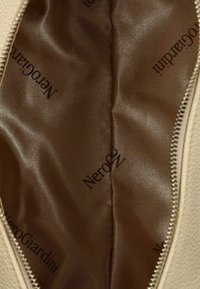 Forro de tela de satén marrón dentro de una bolsa beige con el logo "NeroGiardini" impreso en negro y una cremallera parcialmente visible en el lado derecho.