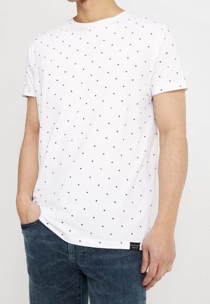 T-shirt en coton blanc à col rond, avec de petits pois bleu marine et rouges, manches courtes et une étiquette à l'ourlet. Associé à un jean foncé.