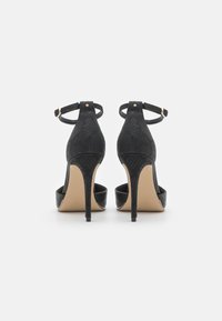 Ein Paar schwarze High-Heels-Sandalen mit Knöchelriemen, strukturiertem Obermaterial und spitzen Stiletto-Absätzen, von hinten auf einem weißen Hintergrund betrachtet.