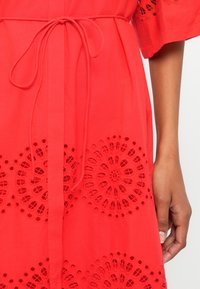 Close-up de um vestido vermelho com recortes de renda circulares, cinto atado na cintura, e o braço e mão direitos de uma pessoa visíveis do lado direito.