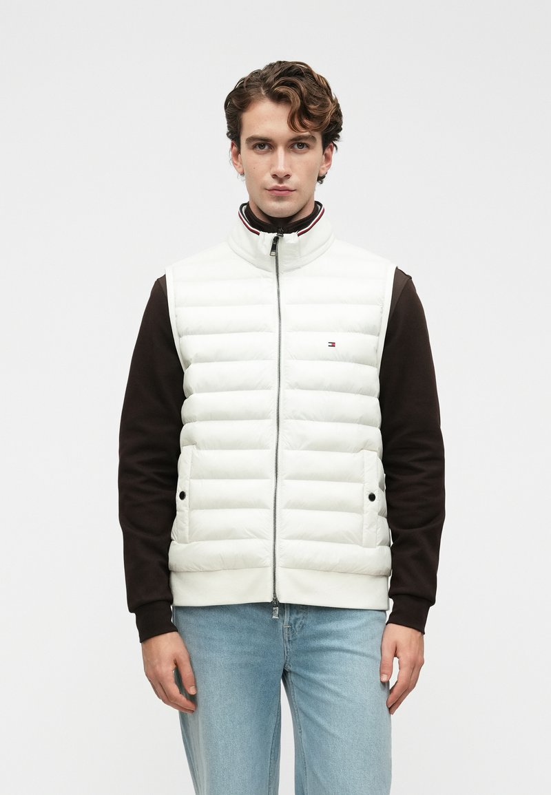 Jaunas vyras su banguotais plaukais, vilkintis šviesiai mėlynus džinsus, tamsią ilgomis rankovėmis marškinį ir baltą siuvinėtą liemenę su priekiu užtrauktuku ir kišenėmis, stovi tiesiai.