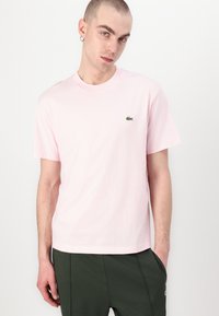 Helles rosa Baumwoll-T-Shirt mit kurzen Ärmeln, Rundhalsausschnitt und kleinem grünen Lacoste-Logo auf der linken Brust. Getragen mit grünen Hosen.
