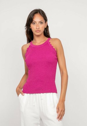 Femme portant un haut en tricot fuchsia sans manches avec des bords festonnés et un pantalon blanc à cordon, debout avec une main dans la poche, sur fond uni.