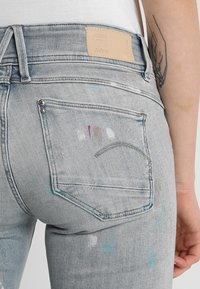 Ljusgrå denimjeans med medelhög midja, med färgsplatter, bakfickor och en märkeslapp i läder på midjan.