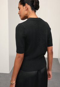 Maglione nero a maniche corte con orlo a coste, caratterizzato da una cucitura posteriore testurizzata. Il materiale sembra morbido e lavorato a maglia.