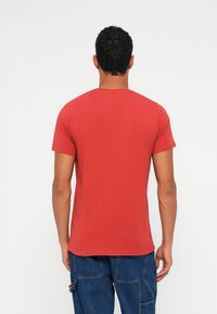 Červené tričko s krátkým rukávem a kulatým výstřihem, měkká textura a přiléhavý střih, zobrazené zezadu na neutrálním pozadí.