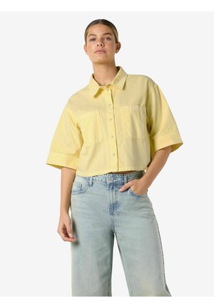 Mladá žena oblečená v žltej krátkoruká košeľa na gombíky a svetlomodré rifle s vysokým pásom, stojaca s jednou rukou v vrecku.