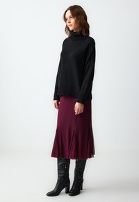 Pull en tricot noir avec col montant, associé à une jupe midi bordeaux à ourlet évasé. Le modèle porte des bottes noires à talons hauts jusqu'aux genoux.