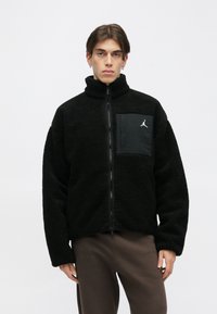 Fekete bolyhos dzseki magas gallérral, cipzáras záródással és logó részletű zsebfelvarrással. Texturált anyag és kényelmes szabás.