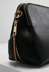 Czarna torba z ekologicznej skóry z złotym suwakiem i elementami metalowymi oraz plecionym, jasnobrązowym dodatkiem przy suwaku, o strukturalnym kształcie i gładkiej powierzchni.