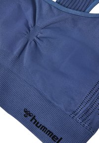 Zbliżenie na niebieski bezszwowy stanik sportowy z marszczonym środkiem i czarnym logo "hummel" na prążkowanym dolnym pasku.