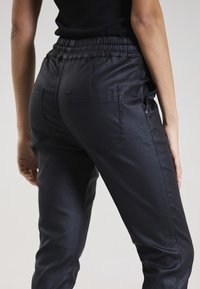 Schwarze, elastische Sweatpants mit glatter, glänzender Oberfläche, ausgestattet mit zwei Gesäßtaschen und einem schmal zulaufenden Bein-Design.