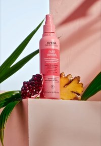 Bouteille de spray rose étiquetée "nutri plenish" avec un bouchon pompe, placée à côté d'une grenade et d'un ananas sur un piédestal rose texturé.