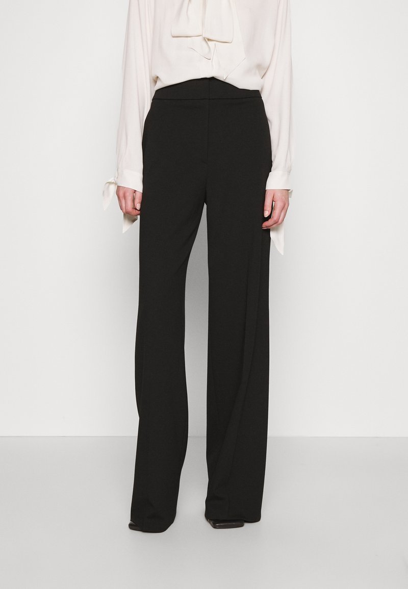 Femme portant un pantalon noir taille haute à jambes larges, associé à un chemisier blanc à manches longues avec des poignets noués.