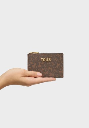 Mano che tiene una piccola pochette marrone con cerniera, con motivi scuri e la scritta dorata "TOUS" sul davanti, su uno sfondo chiaro.