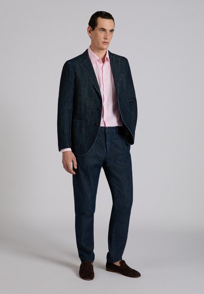 Man standing wearing dark denim suit, light pink button-up shirt, and dark brown loafers against plain gray background.