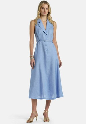 Blondynka w sukience midi niebieskiego koloru z paskiem, bez rękawów, zapinanej na guziki, nosząca złote sandały na obcasie z paseczkami, stojąca na białym tle.