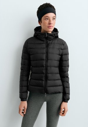 Jeune personne portant une doudoune noire avec capuche, des leggings gris et un bandeau noir, debout devant un fond clair uni.