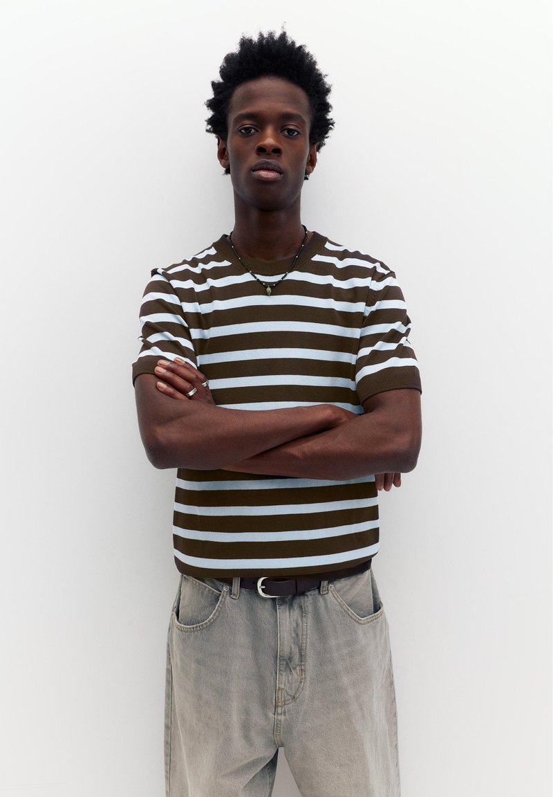 Jeune homme les bras croisés portant une chemise à manches courtes rayée marron et bleu clair, et un jean délavé, debout devant un fond blanc uni.