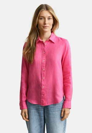 Femme aux longs cheveux blonds portant une chemise rose vif à boutons et un jean bleu clair, debout devant un fond blanc uni.