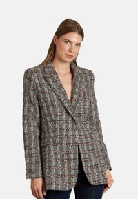 Blazer de tweed gris con un patrón de cuadros, solapas de pico, dos bolsillos y un ajuste ligeramente oversize. Presenta acentos en blanco y marrón por todo el diseño.