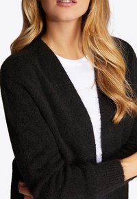 Cardigan nero con texture, aperto davanti e maniche lunghe, indossato sopra una camicia bianca. Materiale morbido e peloso, che evidenzia una vestibilità comoda.