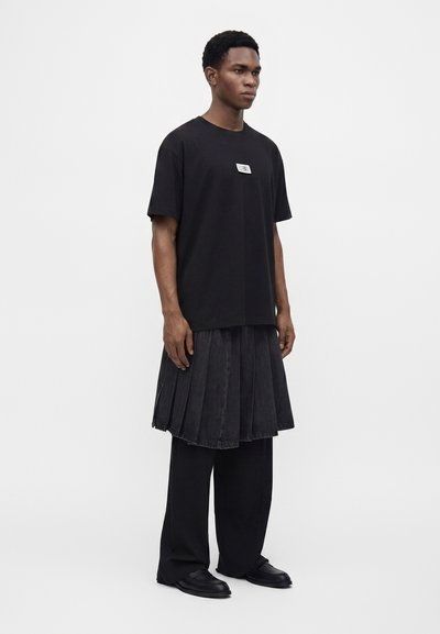 Νέος άνδρας που φοράει μαύρο oversized t-shirt, μαύρη πλισέ φούστα πάνω από μαύρο φαρδύ παντελόνι και μαύρα παπούτσια, στέκεται μπροστά σε απλό φόντο.