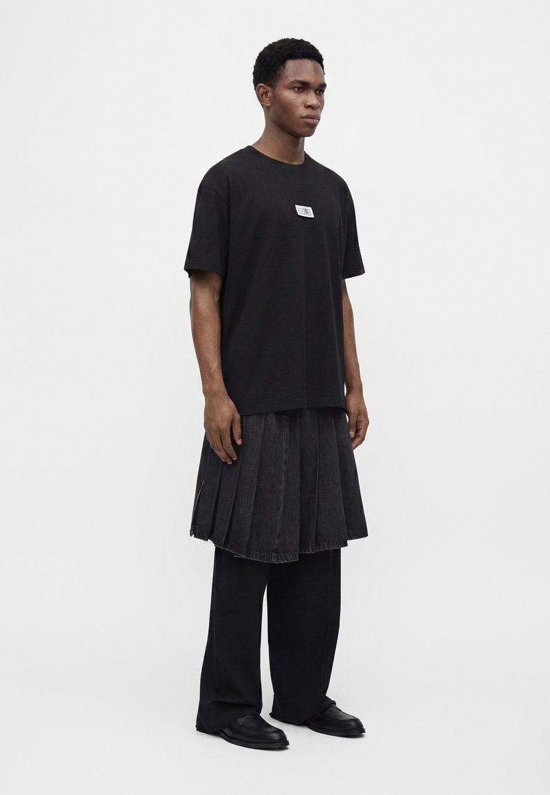 Young man wearing a black oversized t-shirt, a black pleated skirt over black wide pants, and black shoes, standing against a plain background.