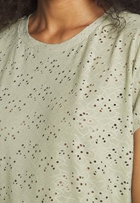 Haut vert clair en tissu texturé avec des motifs découpés complexes et un design de spirale brodé. Manches courtes et décolleté rond.