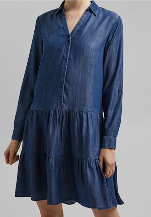 Femme vêtue d'une robe en denim indigo à manches longues, boutonnée, avec une jupe à volants et un col, sur un fond gris uni.