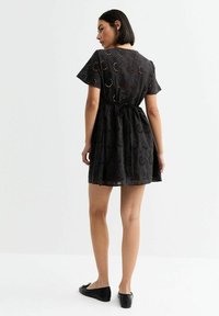 Black, short-sleeved dress with embroidery and cut-out details; cinched waist, flared skirt, and a tie at the back. Flat black shoes.