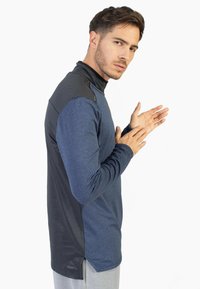 Camiseta atlética de manga larga en tejido azul con un panel texturizado negro en los hombros, que presenta un alto cuello y un dobladillo curvado.