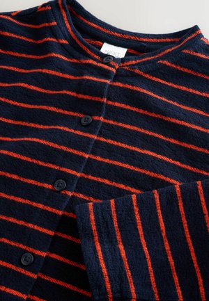 Cardigan bleumarin cu dungi orizontale roșii, realizat dintr-un material moale. Prezintă un decolteu rotund, patru nasturi negri și manșete nervurate.