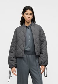 Γκρι μπουφάν με καπιτονέ ραφή, στρογγυλή λαιμόκοψη, κουμπιά με πατιλέτα και ελαστικές μανσέτες. Φορεμένο πάνω από ένα ταιριαστό γκρι σύνολο με διακριτικές λεπτομέρειες λογότυπου.