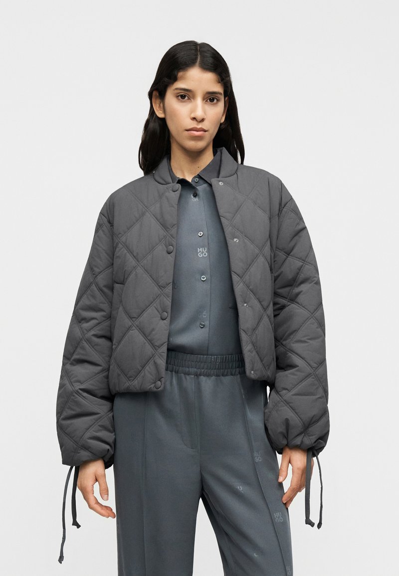 Γκρι μπουφάν με καπιτονέ ραφή, στρογγυλή λαιμόκοψη, κουμπιά με πατιλέτα και ελαστικές μανσέτες. Φορεμένο πάνω από ένα ταιριαστό γκρι σύνολο με διακριτικές λεπτομέρειες λογότυπου.