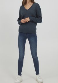 Maglione a collo a V di colore blu navy con maniche lunghe, tessuto testurizzato e orlo a coste, abbinato a jeans skinny blu scuro e sneakers bianche.