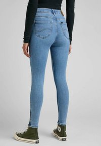 Vaqueros ajustados en denim azul claro, con cintura alta, dos bolsillos traseros y detalles de cremallera en el tobillo, combinados con zapatillas verdes de caña alta.