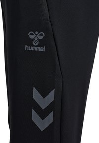 Schwarze Sporthose mit glatter Textur, ausgestattet mit einer Reißverschlusstasche und einem reflektierenden Logo, sowie zwei diagonalen Chevron-Akzenten in Grau.