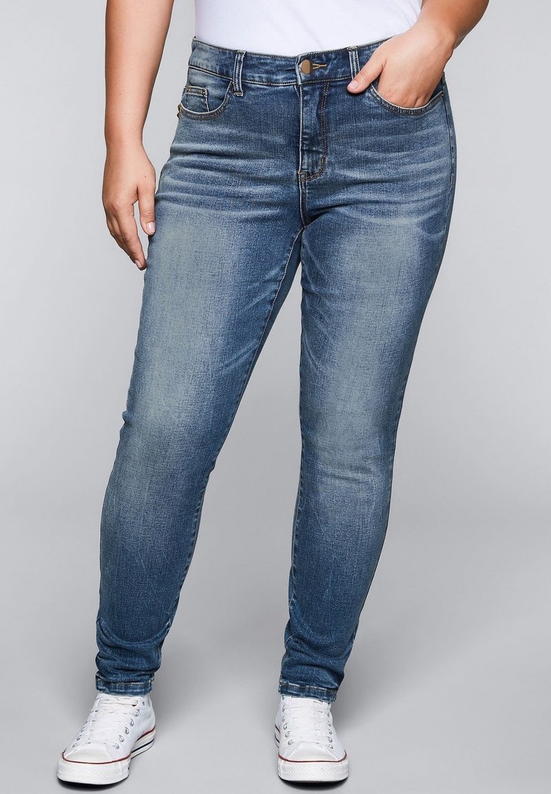 Úzké modré džíny z denimu s vybledlým efektem, pětikapsovým designem a kovovým knoflíkem na zapínání. Kombinovány s bílými teniskami.