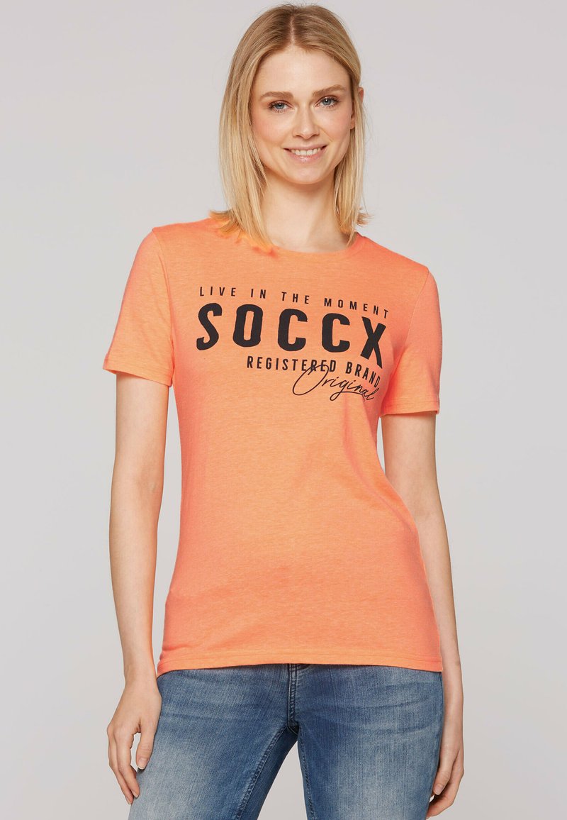 T-shirt arancione a maniche corte realizzato in un tessuto morbido, con una stampa grafica nera e testo. Abbinato a jeans blu, con un semplice scollo rotondo.